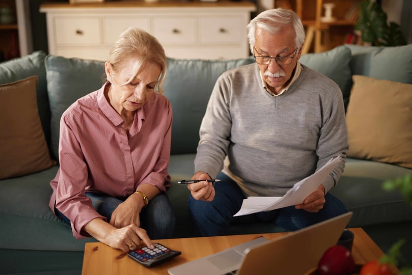 Older man and woman sit on a sofa at home, examining paperwork and using a calculator and a laptop