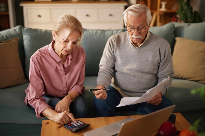 Older man and woman sit on a sofa at home, examining paperwork and using a calculator and a laptop