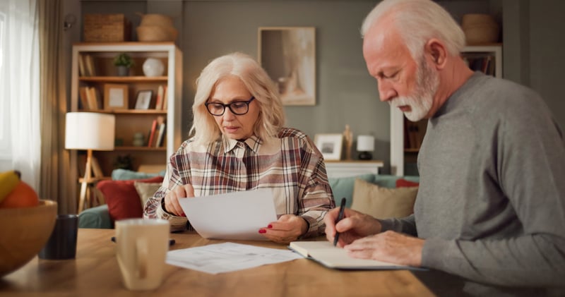 Two older adults examine documents related to life insurance at a table in their living room