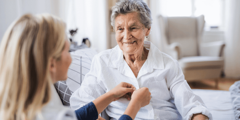 An at-home caregiver helping an older adult woman get dressed. 