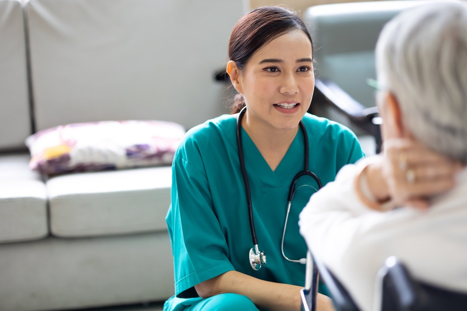 Healthcare professional in teal uniform speaking with an elderly person in a wheelchair.