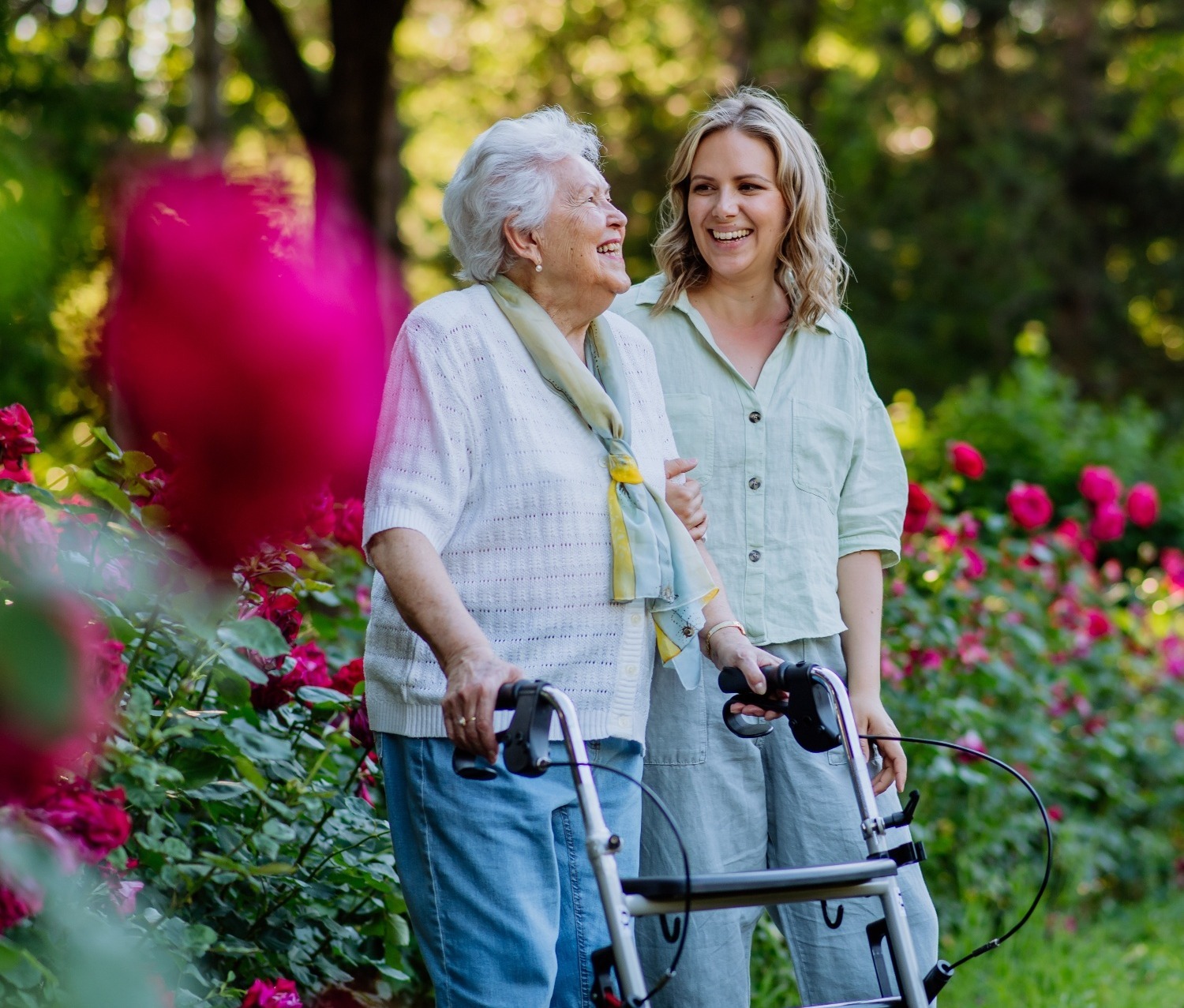 Two women smiling while walking through a garden with red roses, one using a walker.