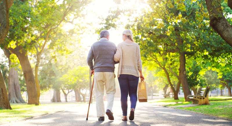 An older man and woman walking arm in arm through a park in the summer.