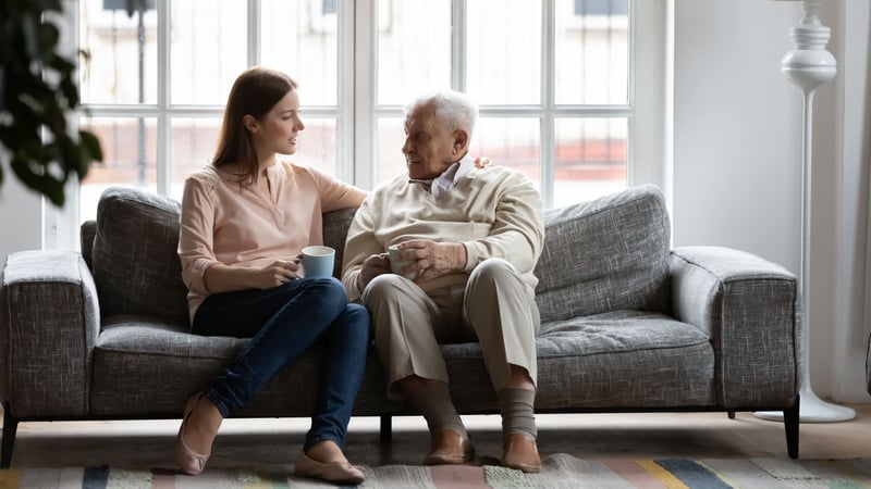 An older man and his adult daughter sitting on a couch in his living room, drinking tea and having a conversation. 