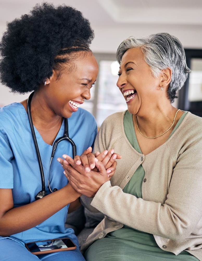 Two women laughing together on a couch, one in medical scrubs and the other in a beige cardigan.