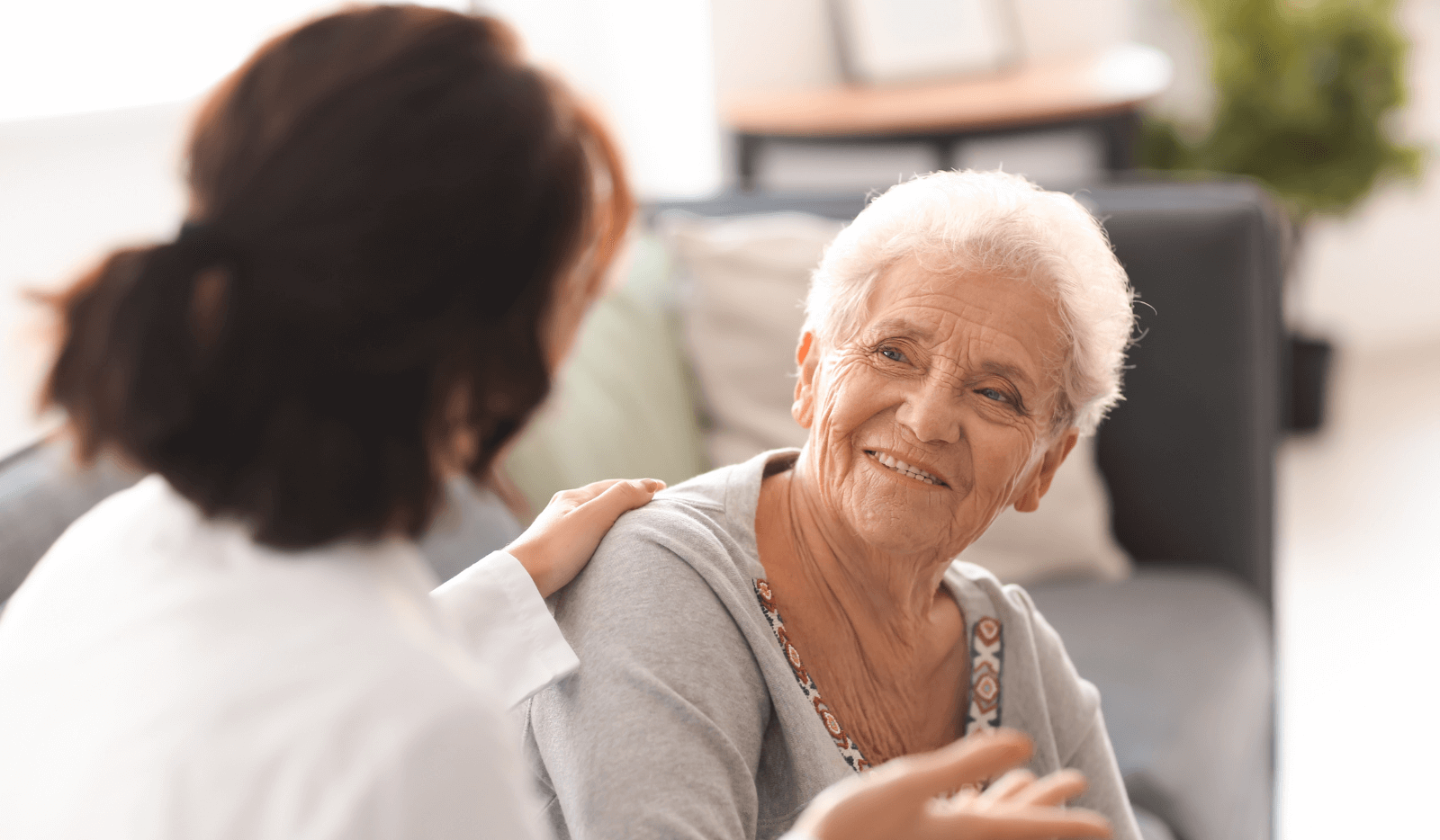 Elderly woman smiling and conversing with another person in a white coat indoors.