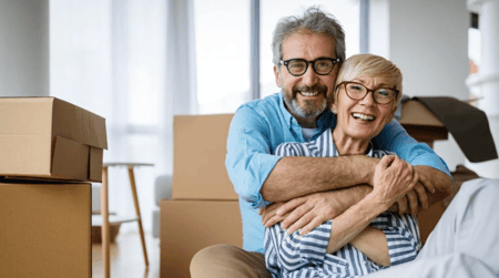 A happy senior couple sitting on the floor surrounded by packed boxes, preparing to downsize their home.