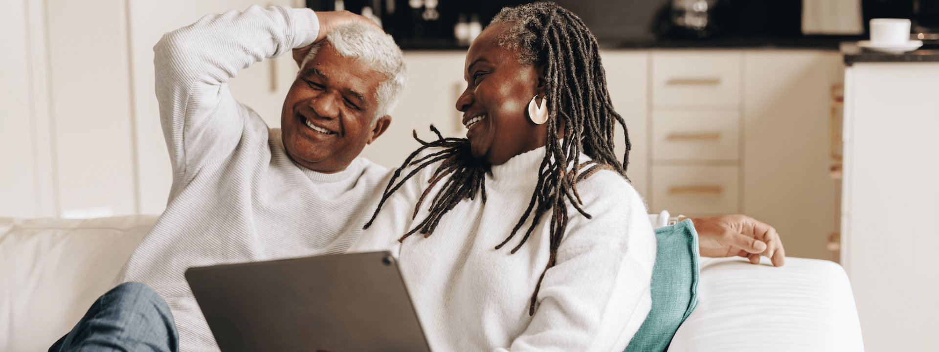 Senior couple sitting on a couch, smiling at each other while holding a tablet.