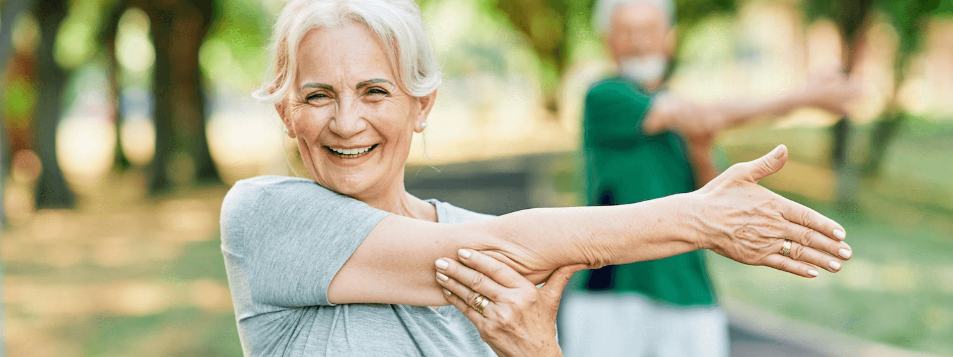 Senior woman smiling while stretching her arm outdoors in a park, with another person stretching in the background.
