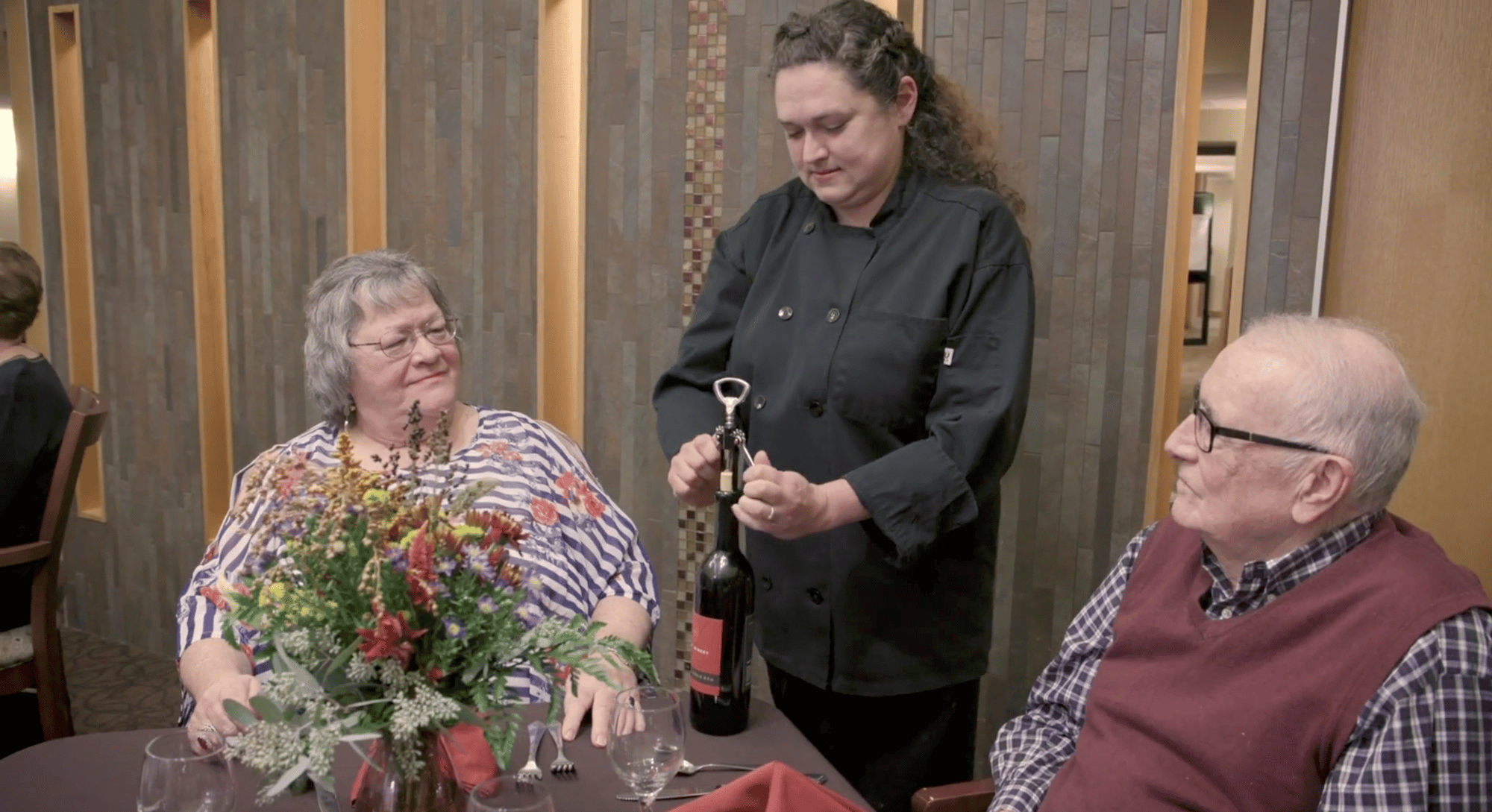Waitress opening a bottle of wine for two older adults