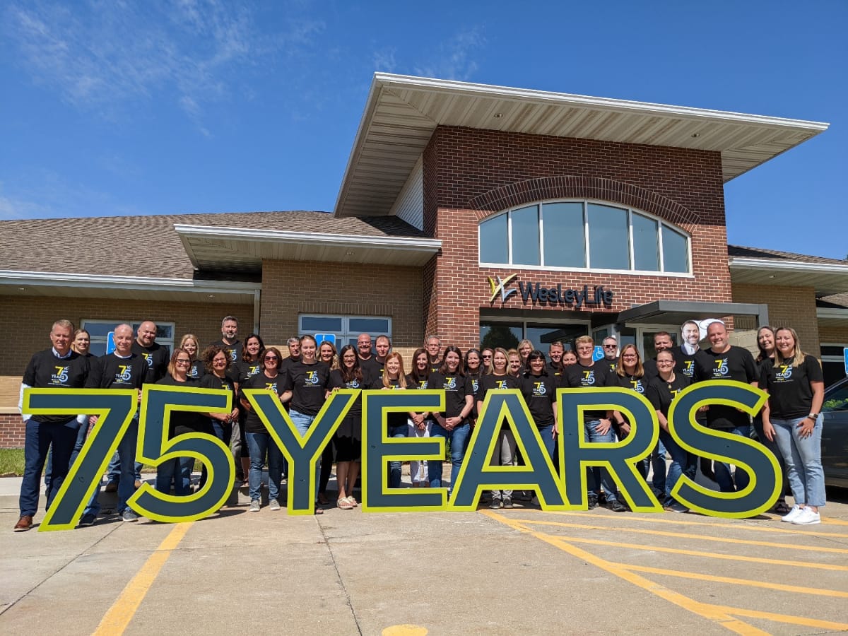 Photo of team standing outside of a WesleyLife building while holding up a sign that reads, 