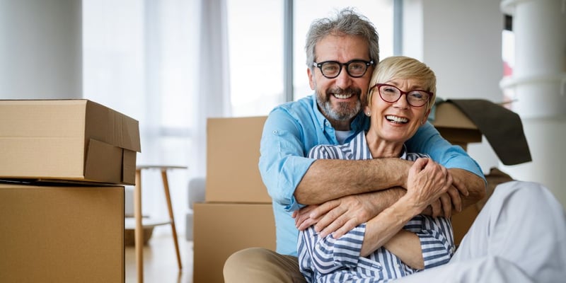 A happy senior couple sitting on the floor surrounded by packed boxes, preparing to downsize their home.