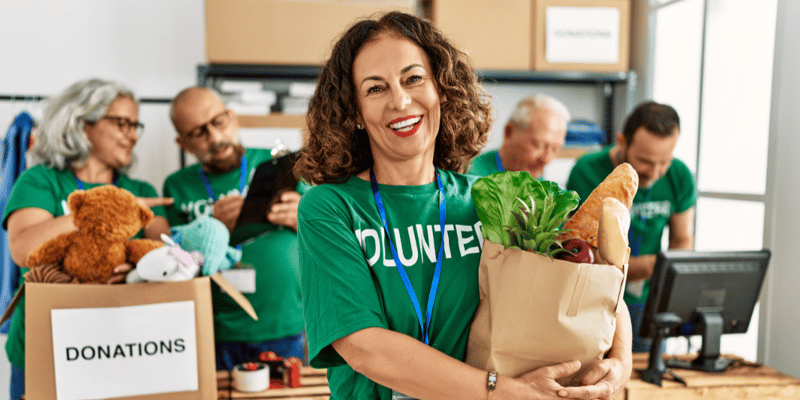A woman wearing a “VOLUNTEER” shirt and holding a bag of produce at a donation station. There are four other volunteers behind her. 