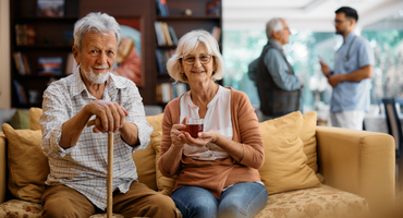 A smiling senior couple sitting on a couch. The man is holding a cane, and the woman has a cup of tea.