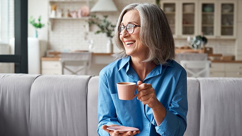 Happy beautiful relaxed mature older adult grey haired woman drinking coffee relaxing on sofa at home