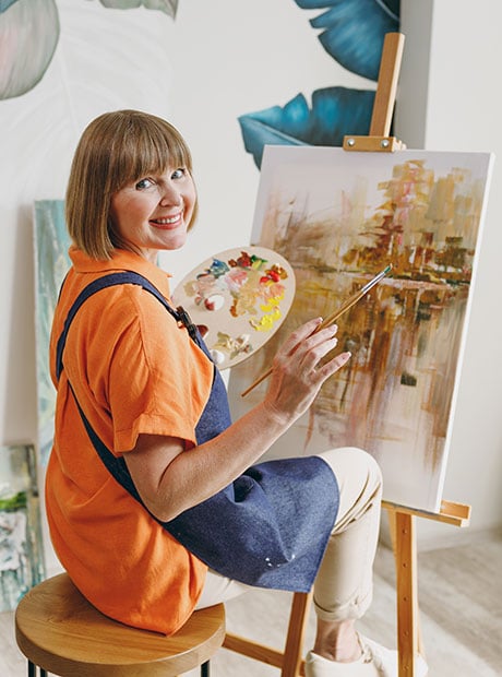 An elderly artist woman wearing casual clothes sitting near an easel with painting artwork painting