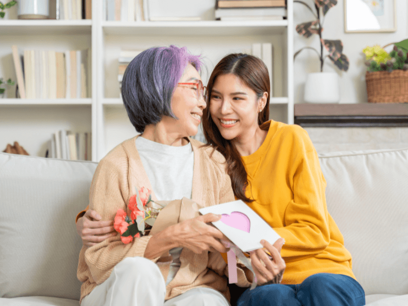 Two women sitting together on a sofa, holding a gift and flowers, smiling at each other lovingly.