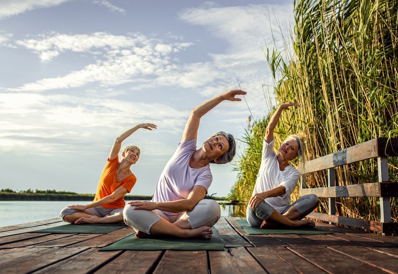 Three senior women practicing yoga on a wooden deck by the water.