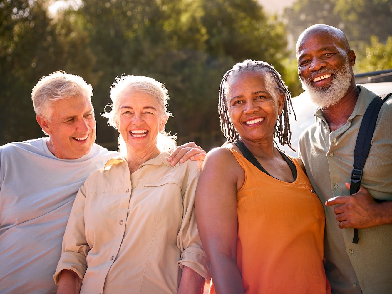 Four adults smiling together outdoors with a sunny, tree-filled background.