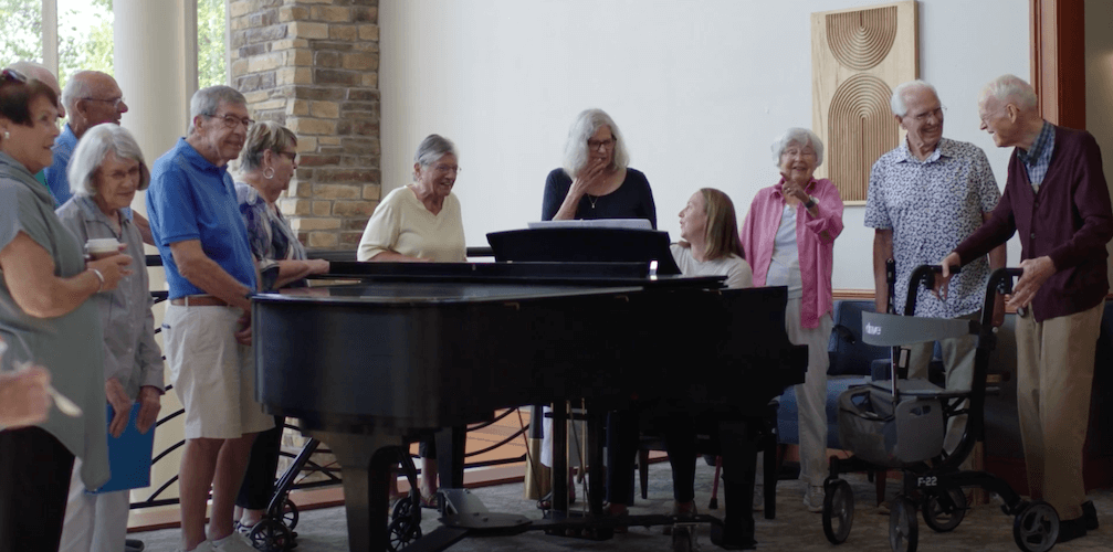 A group of elderly adults around a grand piano in a room with light walls and a stone pillar.