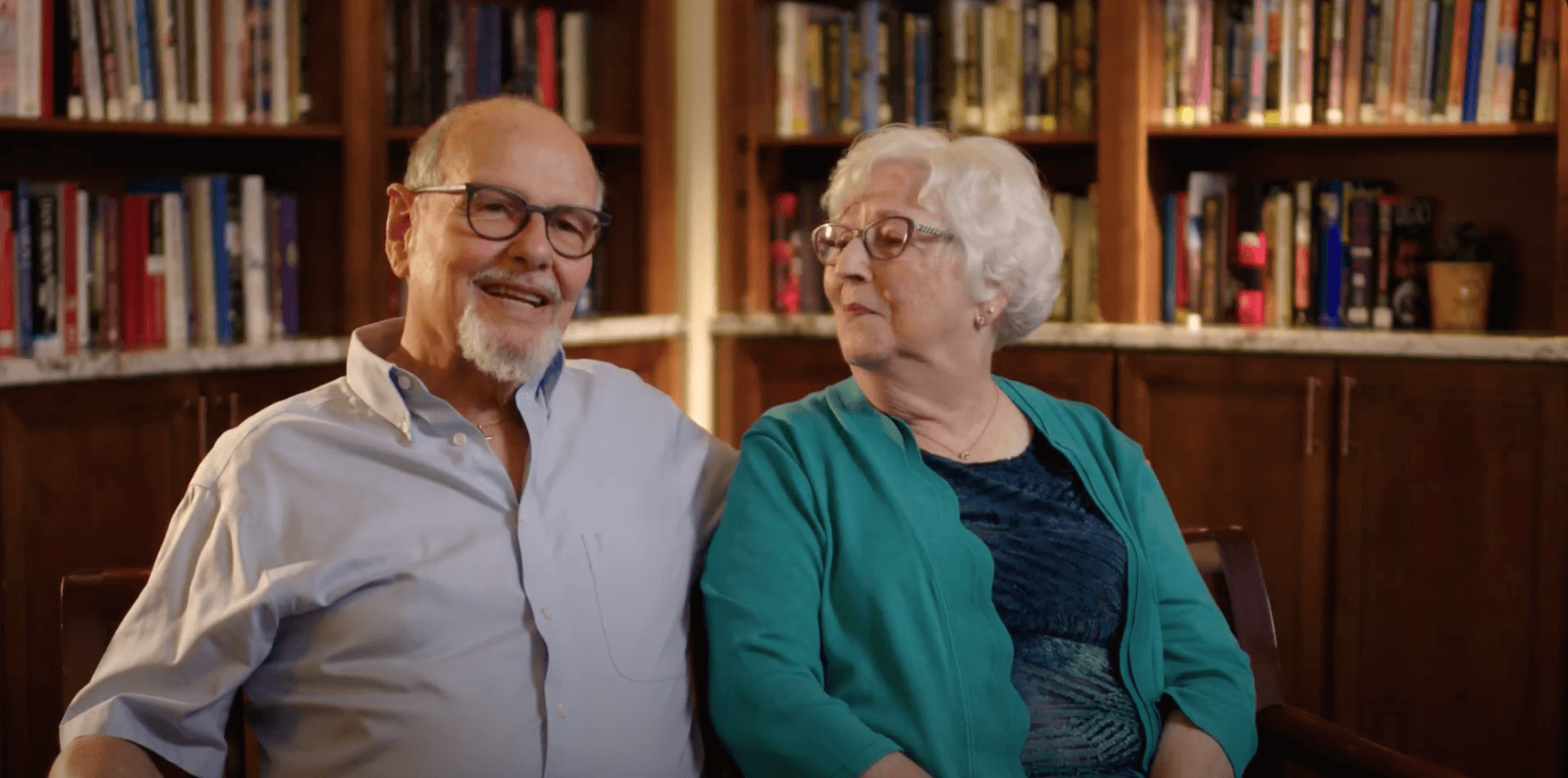 An older couple sitting together in front of bookshelves, the man smiling and the woman looking at him.